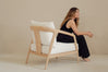 A woman in a black sleeveless outfit sits barefoot on a light wood armchair with white cushions, photographed in a minimalist studio setting with a warm beige background.