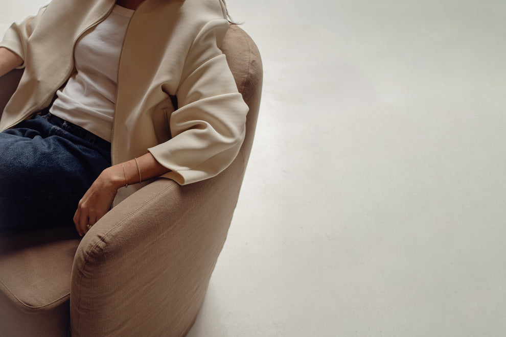 A person seated in a beige upholstered armchair, wearing a cream jacket over a white top and dark blue jeans, photographed in a minimalist indoor setting with a light grey floor.