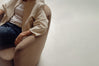 A person seated in a beige upholstered armchair, wearing a cream jacket over a white top and dark blue jeans, photographed in a minimalist indoor setting with a light grey floor.