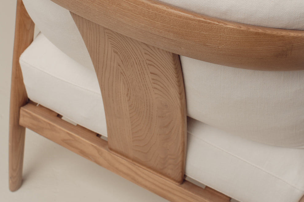 Close-up of a wooden armchair with a light oak frame and off-white cushions, showing the grain and texture of the wood and fabric.