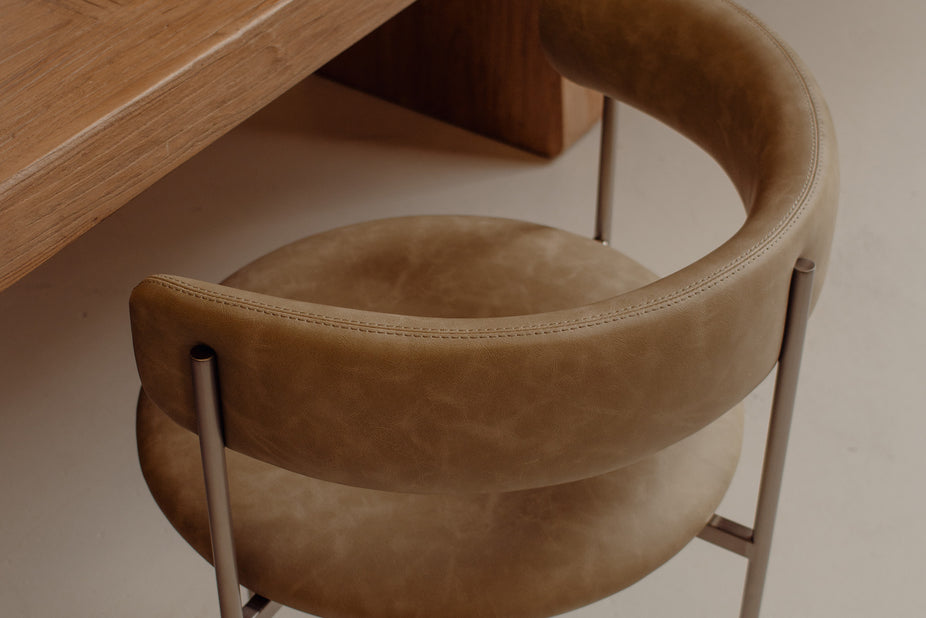 A close-up of a modern curved upholstered chair in warm tan leather with slim metallic legs, shown in a minimalist interior setting next to a wooden table.