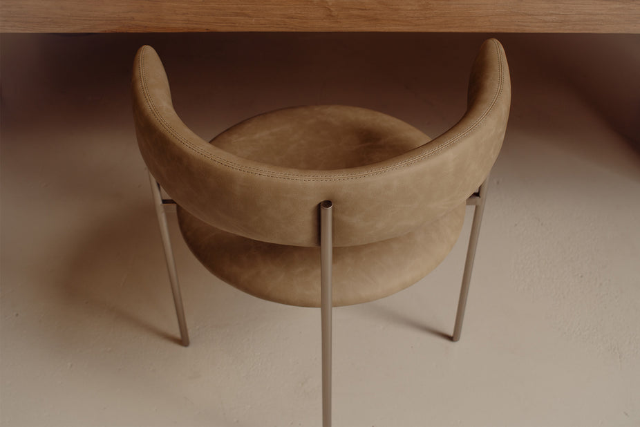 A modern upholstered accent chair with a curved backrest and seat, upholstered in a warm taupe-brown suede-like material, supported by slim metallic legs, shown in a minimalist interior setting.