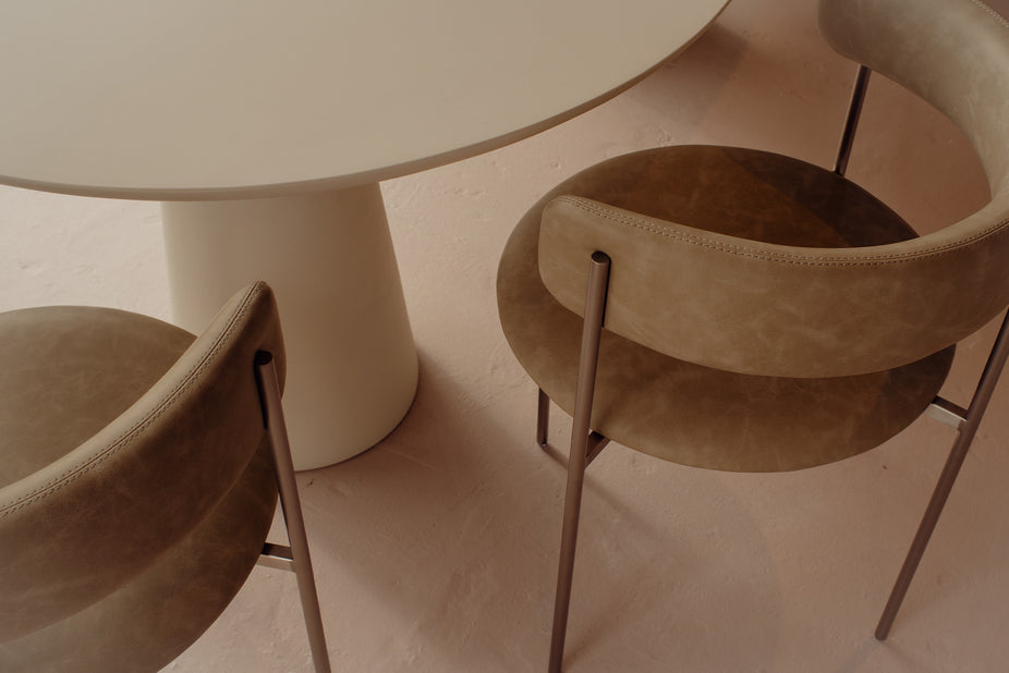 A close-up view of two modern upholstered dining chairs with a warm taupe-brown vinyl finish and slim metal legs, positioned around a round table on a matching beige floor.