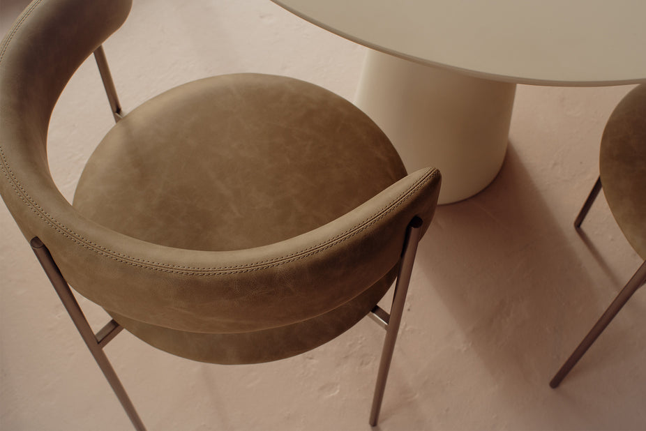 A close-up, high-angle view of a modern upholstered dining chair in a warm taupe-brown vinyl fabric with a curved backrest and slim metal legs, positioned next to a white table.