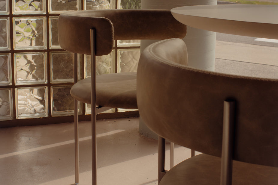 A close-up of a modern curved upholstered dining chair in a warm taupe-brown velvet or suede-like material with slim metallic legs, set in a softly lit interior near a glass block wall and table.