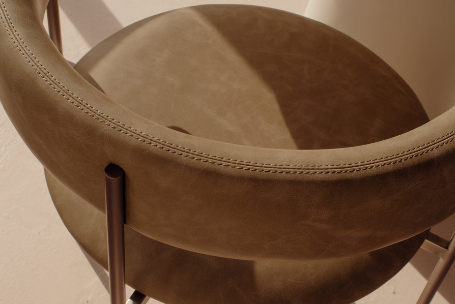 Close-up of a modern upholstered chair with a curved backrest and seat, upholstered in taupe-brown leather or faux leather, featuring visible stitching and dark metal legs.