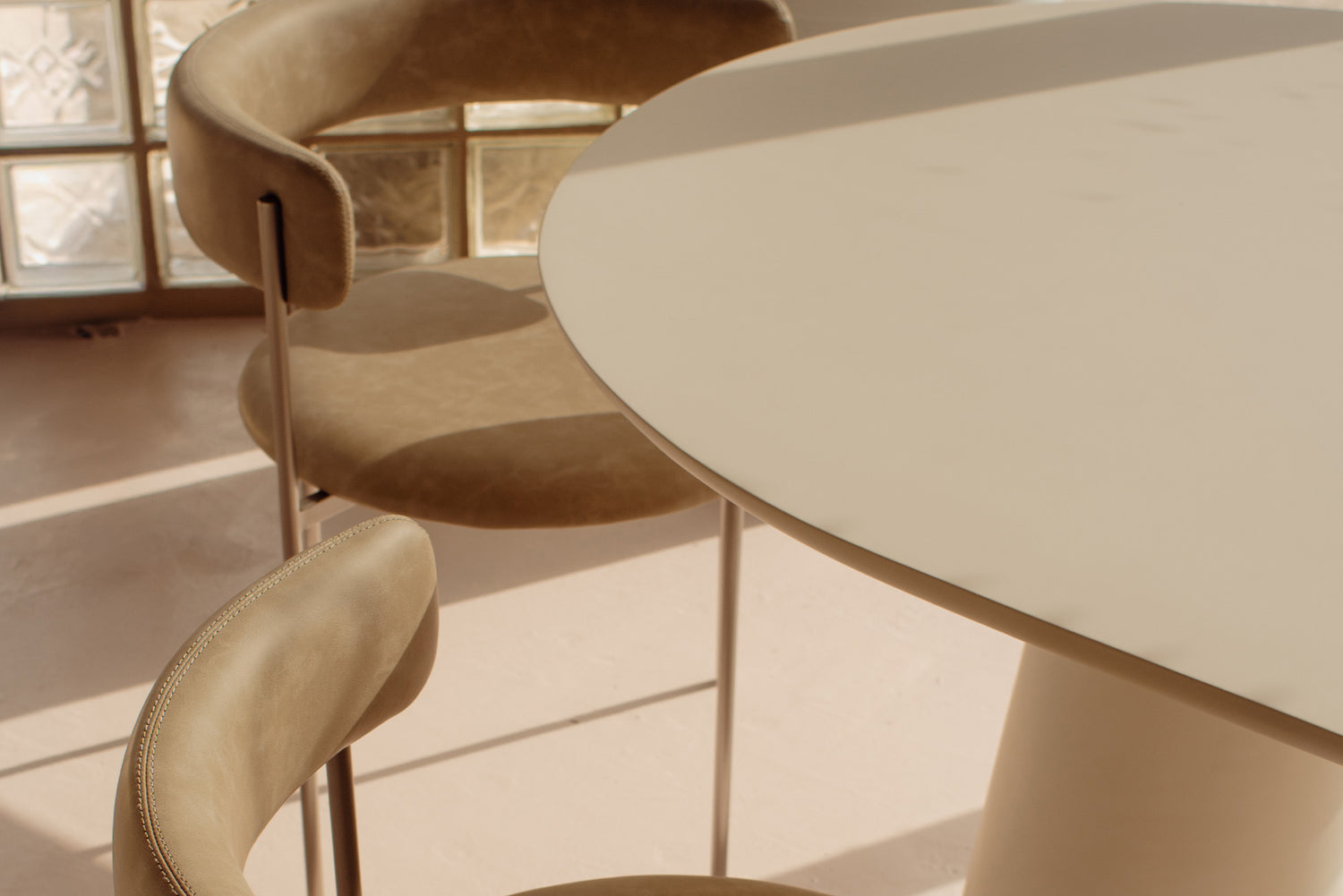 A close-up of a modern dining setup featuring a round off-white table and two upholstered bar stools in a warm beige tone, set in a brightly lit room with natural light.