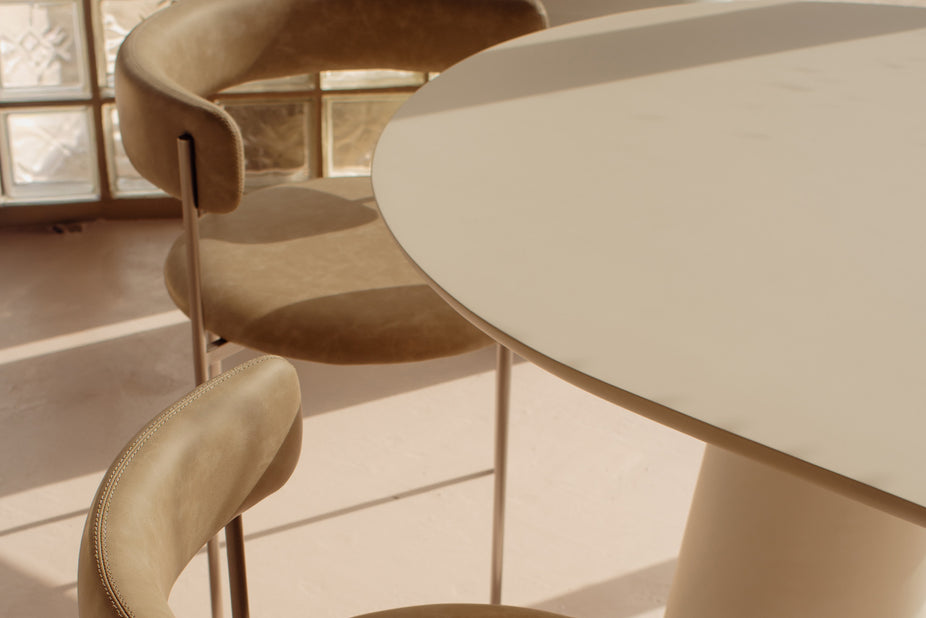 A close-up of a modern dining setup featuring a round off-white table and two upholstered bar stools in a warm beige tone, set in a brightly lit room with natural light.