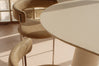A close-up of a modern dining setup featuring a round off-white table and two upholstered bar stools in a warm beige tone, set in a brightly lit room with natural light.