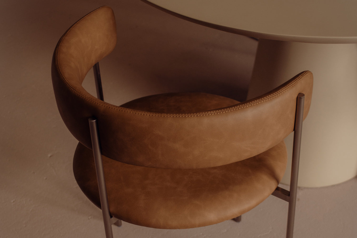 A close-up of a modern dining chair with a curved back and seat in warm brown leather, supported by slim dark metal legs, set in a minimalist interior next to a round table.