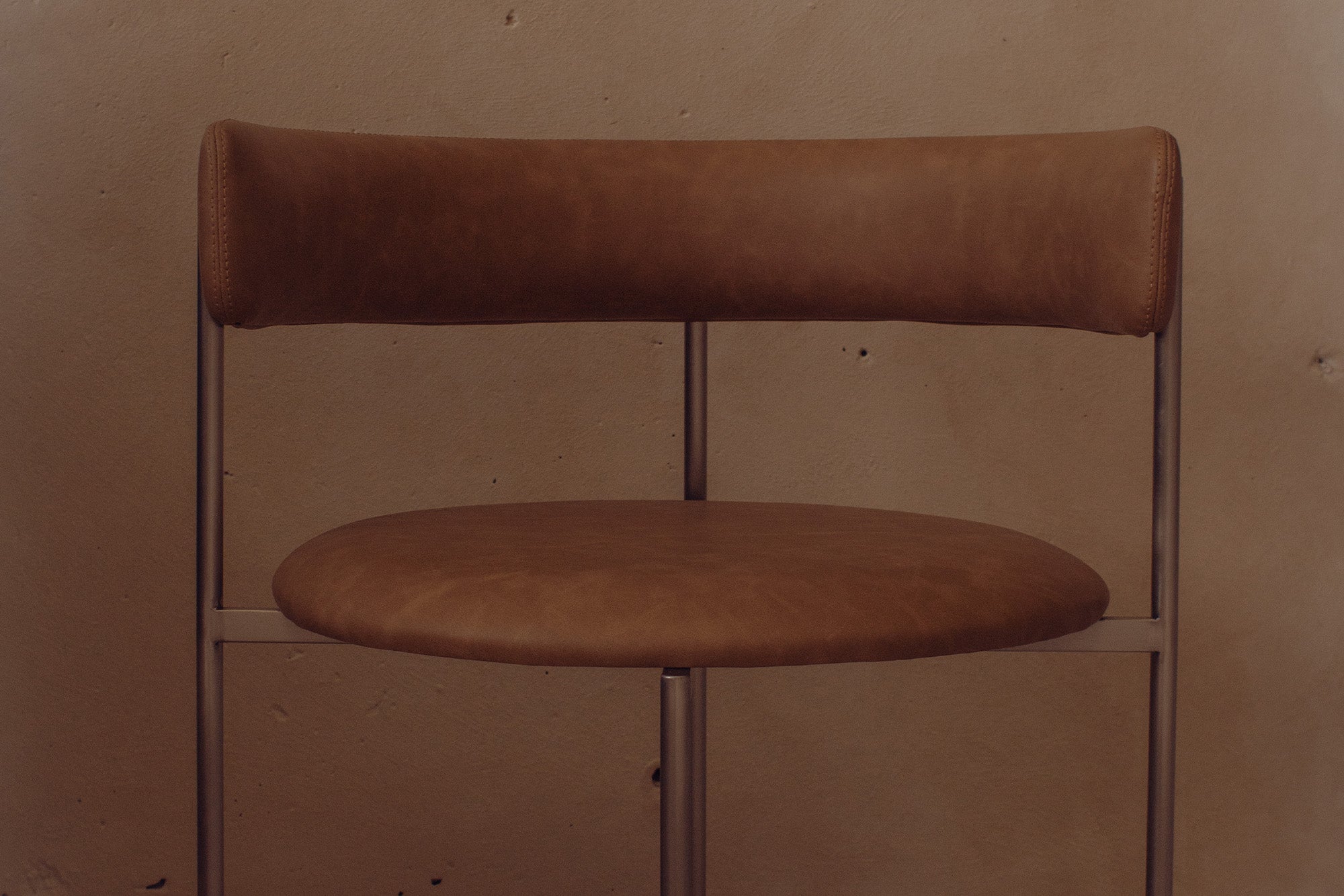 A close-up of a modern dining chair with a curved back and seat in warm brown leather, supported by slim dark metal legs.