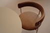 A mid-century modern accent chair with a curved brown leather backrest and seat on slim metal legs, shown next to a matching round table against a neutral beige wall.