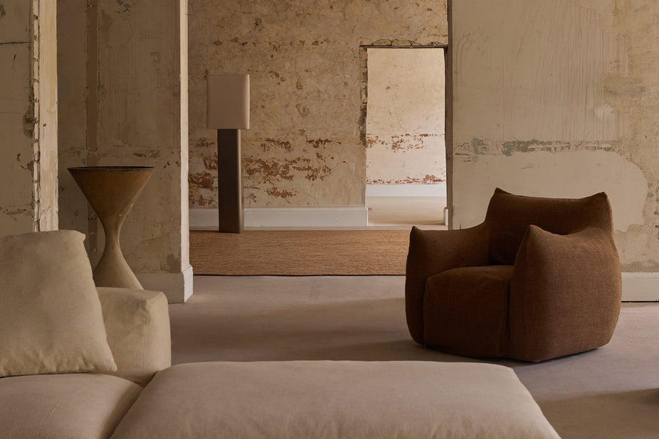 A modern, low-profile armchair upholstered in a rich brown fabric, styled in a minimalist living room with textured plaster walls and neutral-toned furniture.