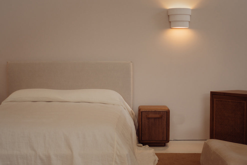 A minimalist bedroom scene featuring a bed with white linens and a beige upholstered headboard, a small wooden nightstand, a wall-mounted cylindrical sconce, and a wooden cabinet against a warm beige wall.
