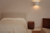 A minimalist bedroom scene featuring a bed with white linens and a beige upholstered headboard, a small wooden nightstand, a wall-mounted cylindrical sconce, and a wooden cabinet against a warm beige wall.