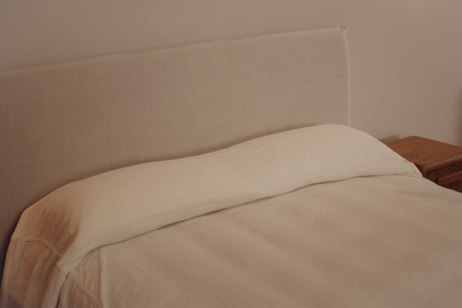 A close-up of a bed with a cream-colored upholstered headboard, a matching cream pillow, and light beige bedding, next to a wooden bedside table.