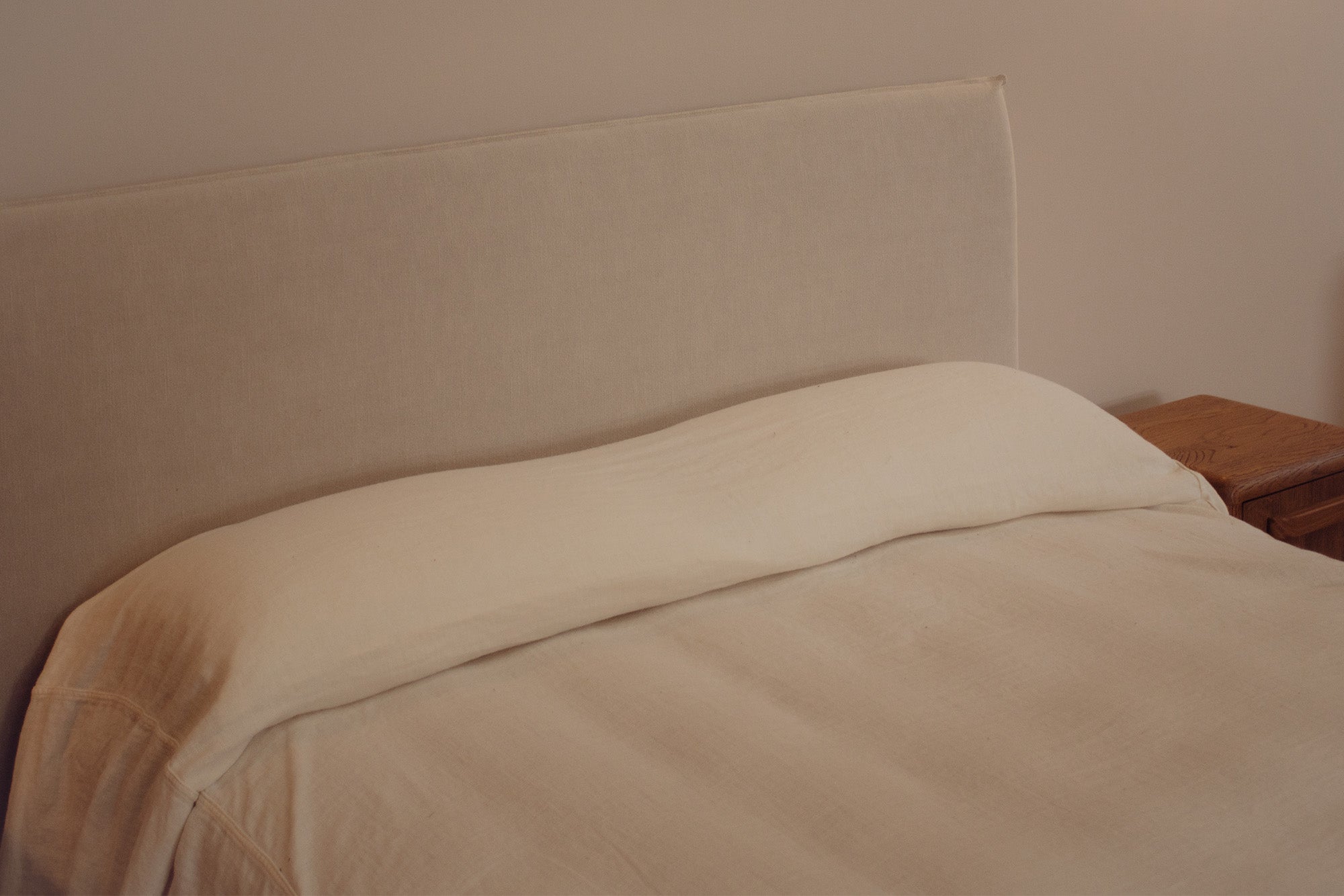 A close-up of a bed with a cream-colored upholstered headboard, a matching cream pillow, and light beige bedding, next to a wooden bedside table.