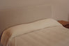 A close-up of a bed with a cream-colored upholstered headboard, a matching cream pillow, and light beige bedding, next to a wooden bedside table.