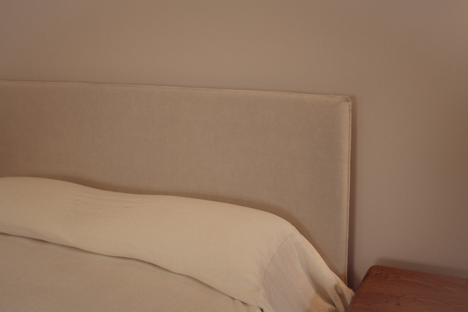 Beige upholstered bed headboard with a cream pillow and white bedding, shown in a minimalist bedroom setting next to a wooden nightstand.