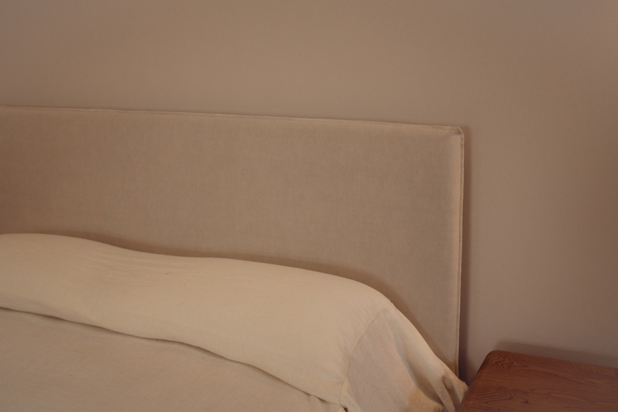 Beige upholstered bed headboard with a cream pillow and white bedding, shown in a minimalist bedroom setting next to a wooden nightstand.