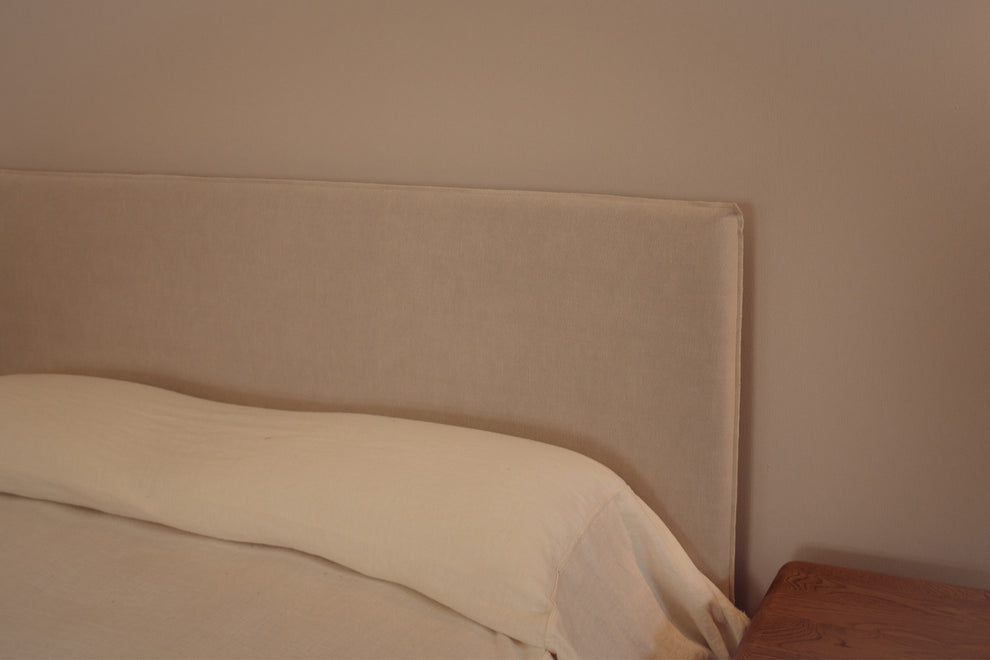 Beige upholstered bed headboard with a cream pillow and white bedding, shown in a minimalist bedroom setting next to a wooden nightstand.