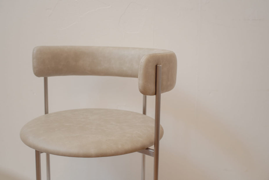 A modern bar stool with a round upholstered seat and a curved backrest, upholstered in a light beige textured fabric with slim metallic legs, set against a plain off-white wall.