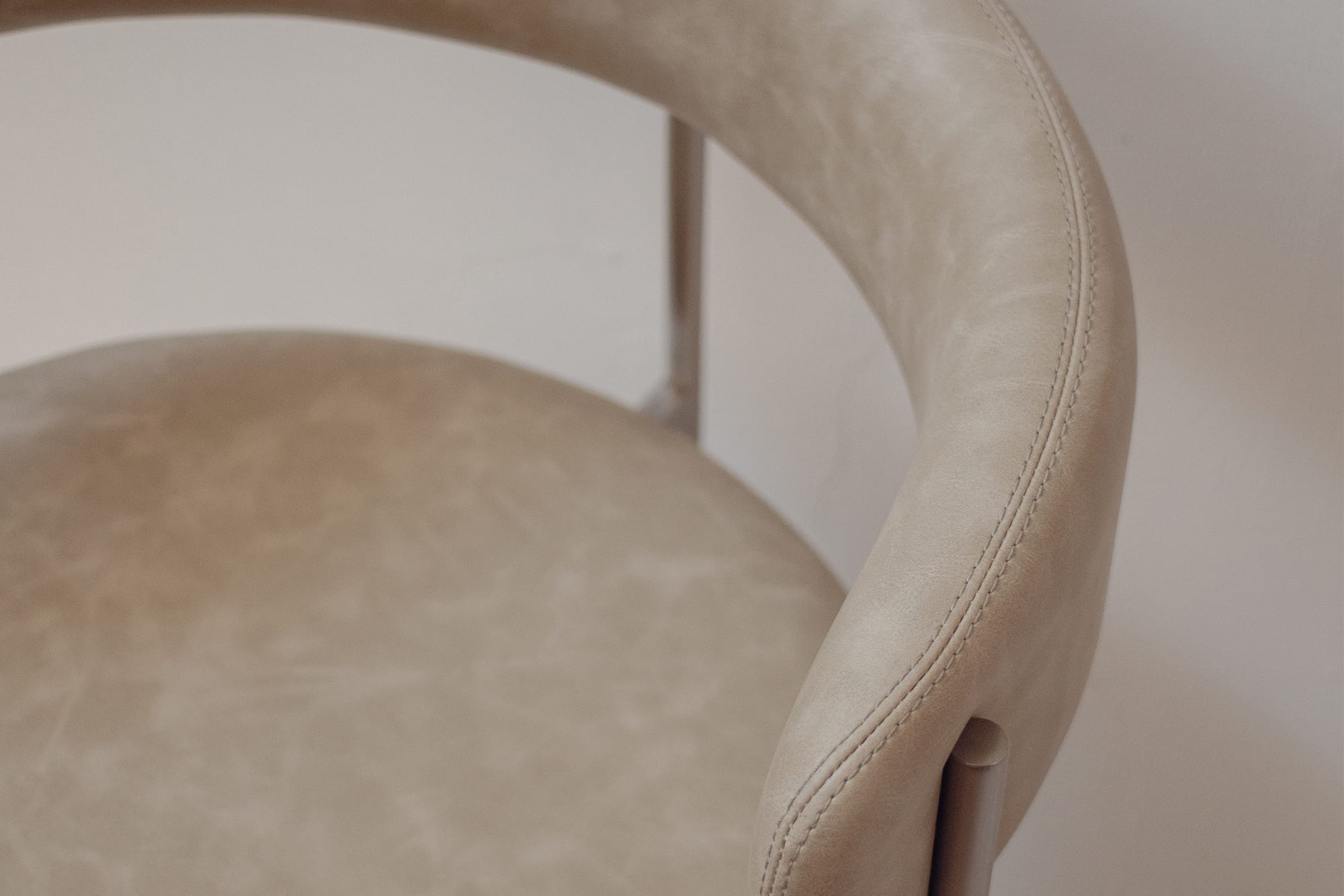 Close-up of a modern upholstered chair with a curved backrest and seat in a light beige or taupe leather-like material, showing a slim metallic support leg against a plain light background.