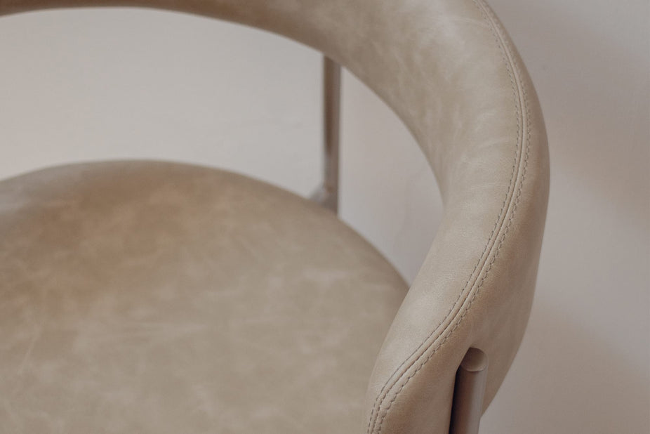 Close-up of a modern upholstered chair with a curved backrest and seat in a light beige or taupe leather-like material, showing a slim metallic support leg against a plain light background.