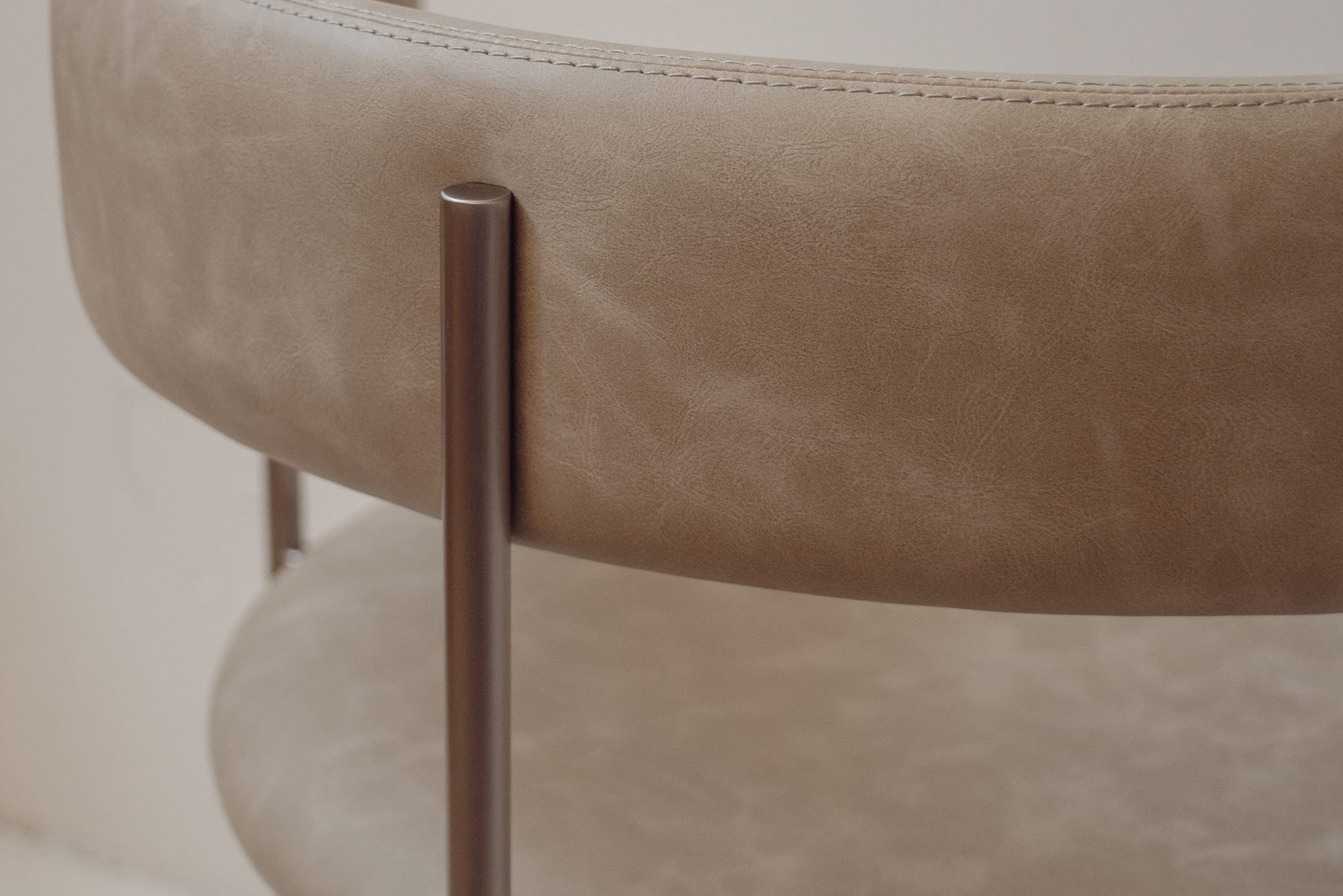 Close-up of a modern upholstered chair with a curved taupe leather seat and back, featuring a brushed metal cylindrical leg.