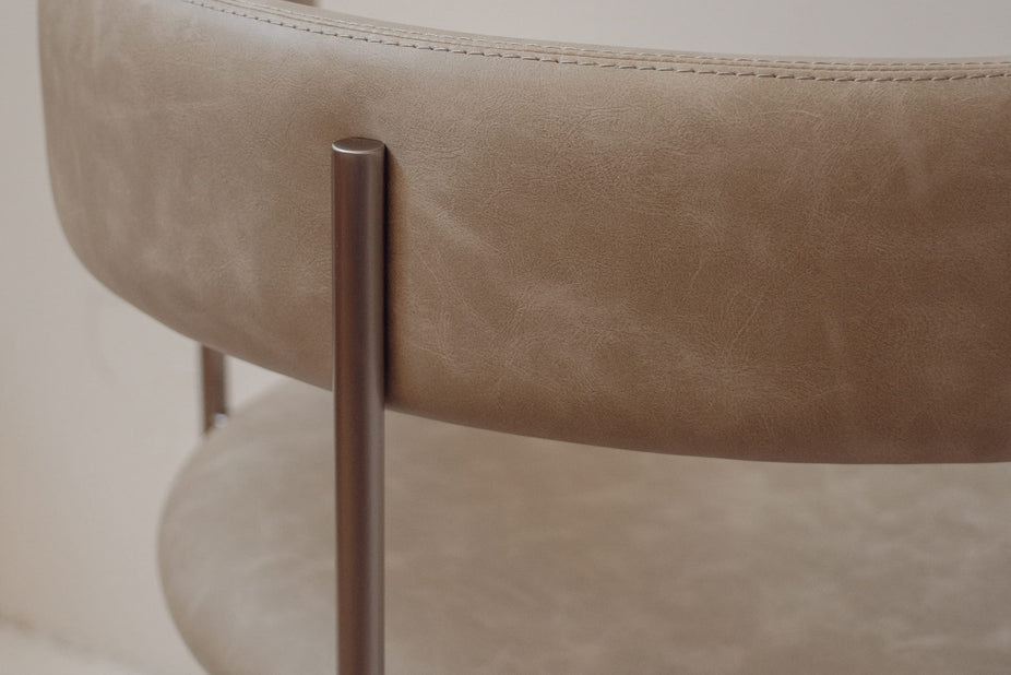 Close-up of a modern upholstered chair with a curved taupe leather seat and back, featuring a brushed metal cylindrical leg.
