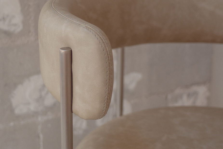 Close-up of a modern upholstered chair with a rounded beige leather backrest and a slim metallic silver frame, set against a textured gray plaster wall.