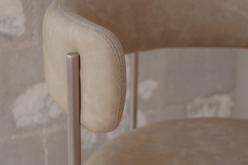 Close-up of a modern upholstered chair with a rounded beige leather backrest and a slim metallic silver frame, set against a textured gray plaster wall.