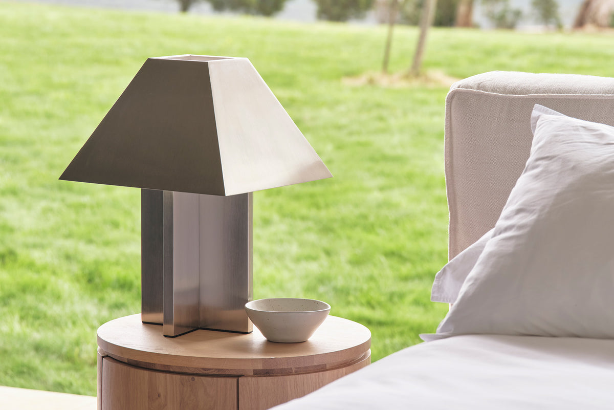 A brushed steel table lamp with a geometric design with a grassy field in the background.