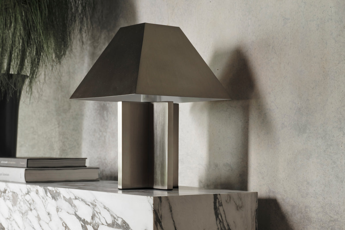 A brushed steel table lamp with a geometric design, placed on a marble surface, with a plant in the background.